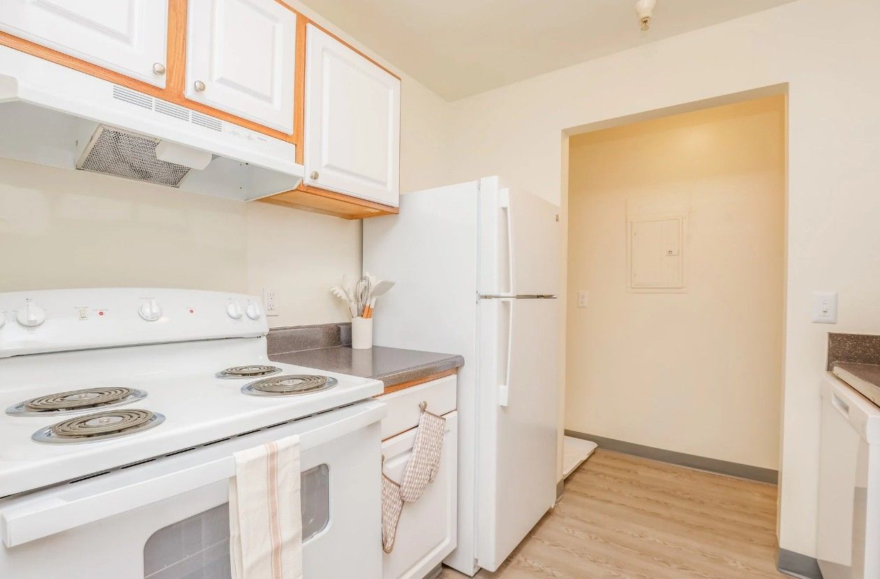 A clean kitchen with white appliances, wooden cabinets, a stove, and wood-look flooring leading to an open doorway.