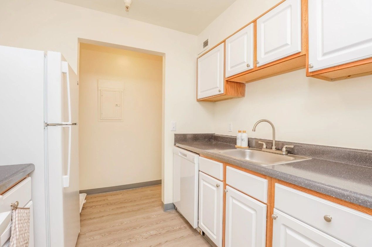 A small, brightly lit kitchen featuring white cabinets, gray countertops, a stainless steel sink, and a white refrigerator.
