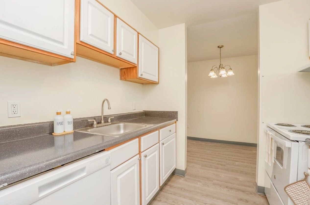 A clean kitchen featuring white cabinets, grey countertops, a sink, and a white stove, opening to a small dining area.