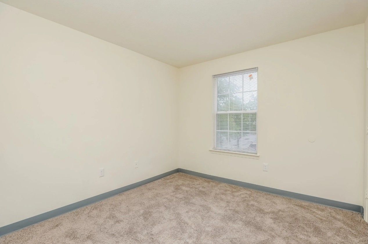 An empty room with cream walls, beige patterned carpeting, and a single window with white trim.