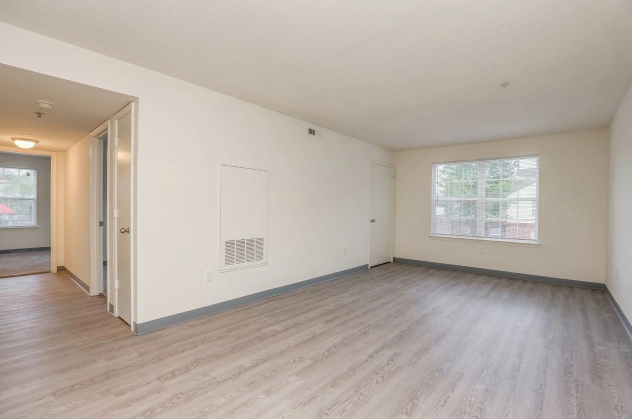 An empty, modern apartment living room with light wood-style flooring, white walls, and a large window facing outdoors.
