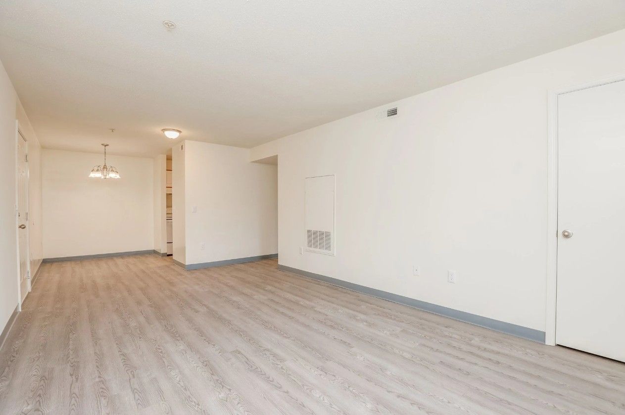 A wide-angle interior view of an empty living area with light-toned wood flooring, white walls, and a closed white door.
