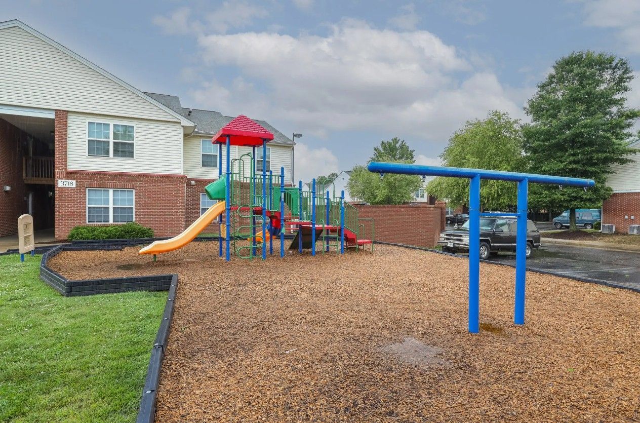 A playground with a colorful play structure and slide set in wood mulch next to a brick and siding apartment building.