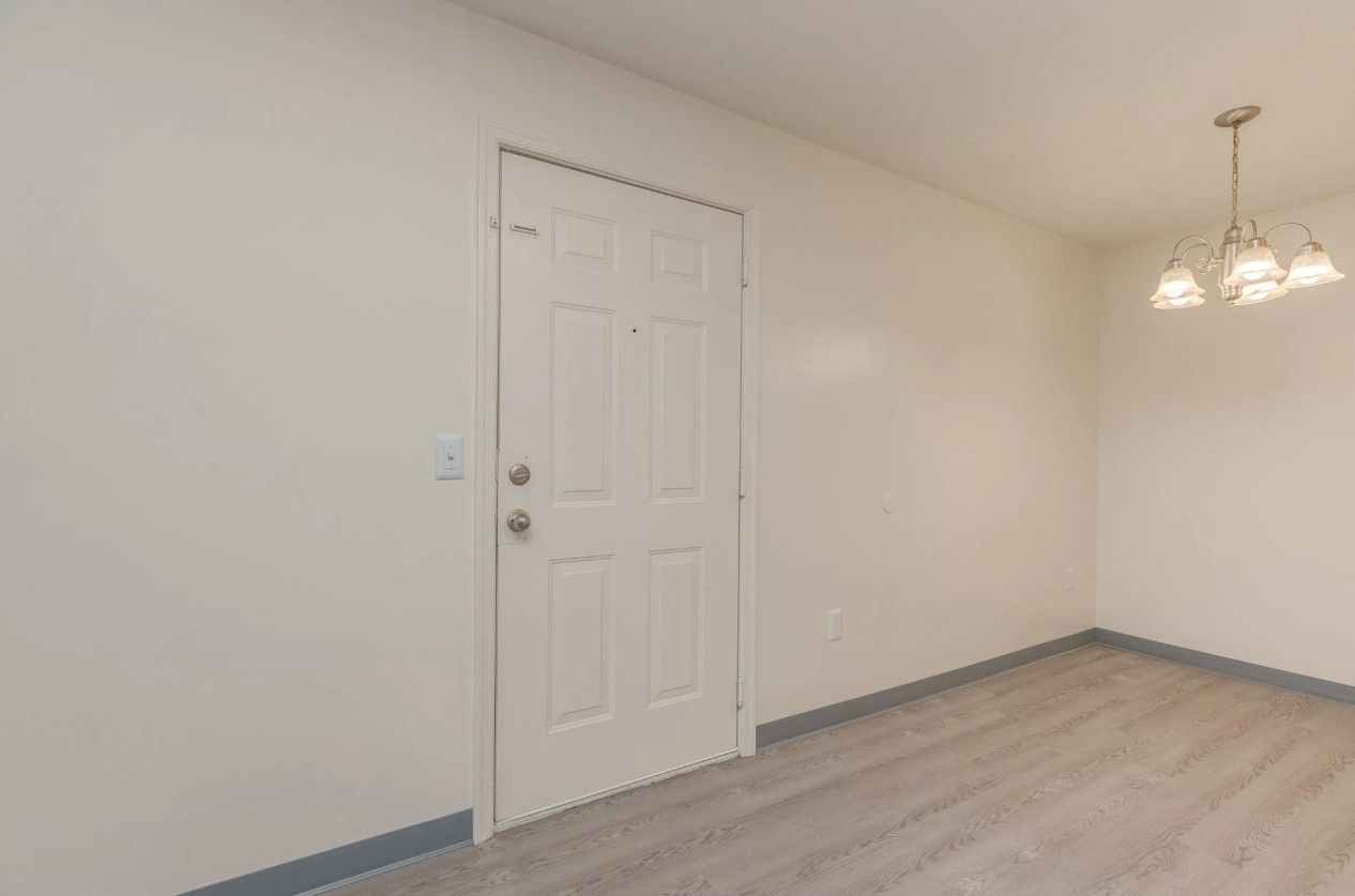An interior view of a room with light grey wood flooring, plain white walls, a white front door, and a hanging chandelier.