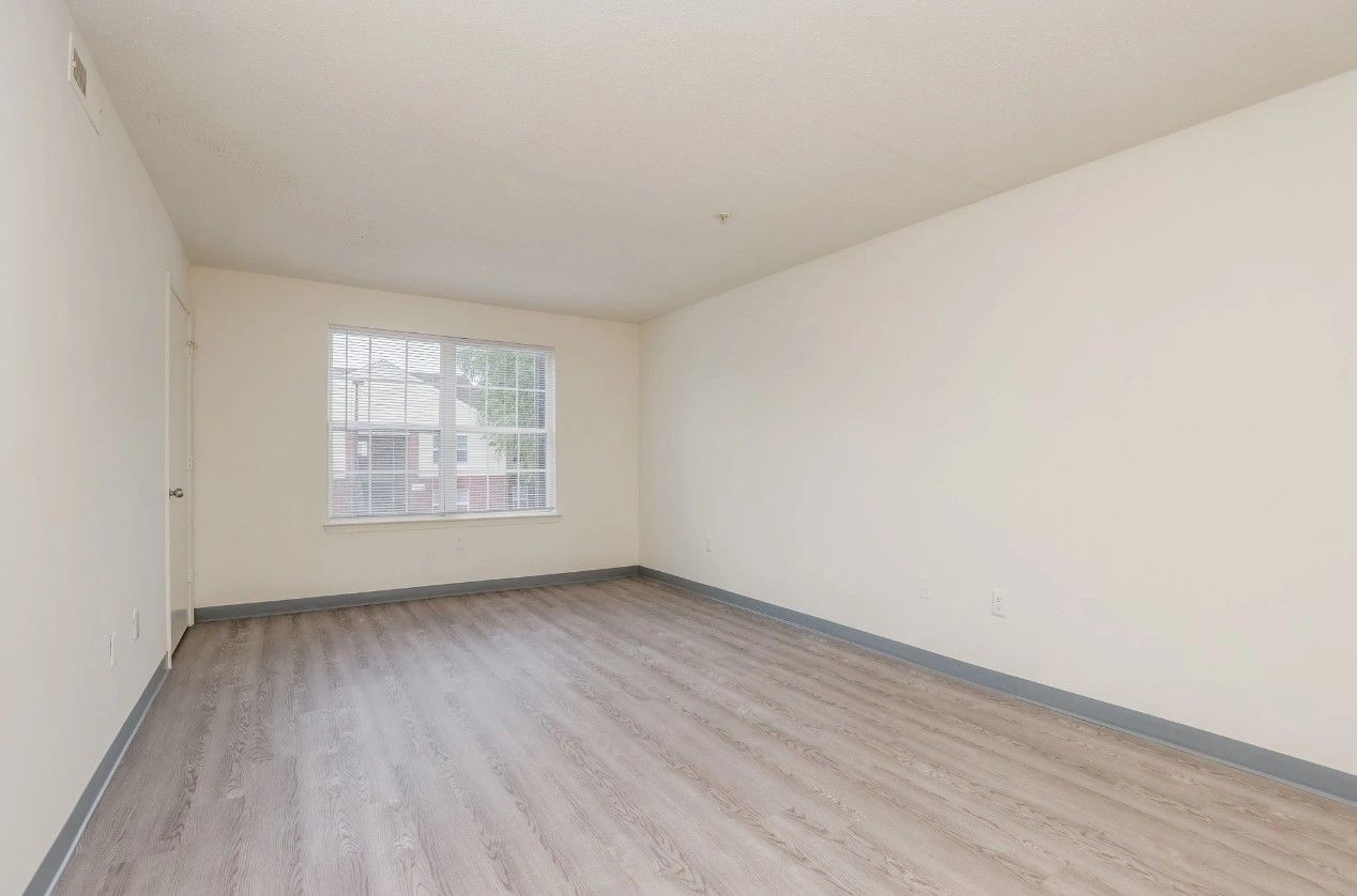An empty room with light wood-look flooring, cream-colored walls, and a large window.
