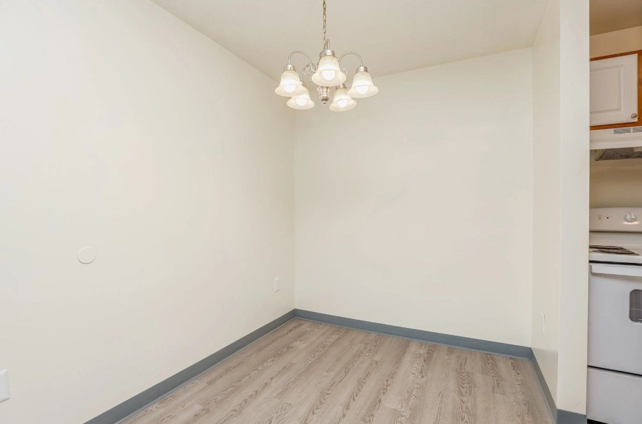 An empty dining area with light walls, wood-look flooring, a five-light chandelier, and a partial view of a kitchen.