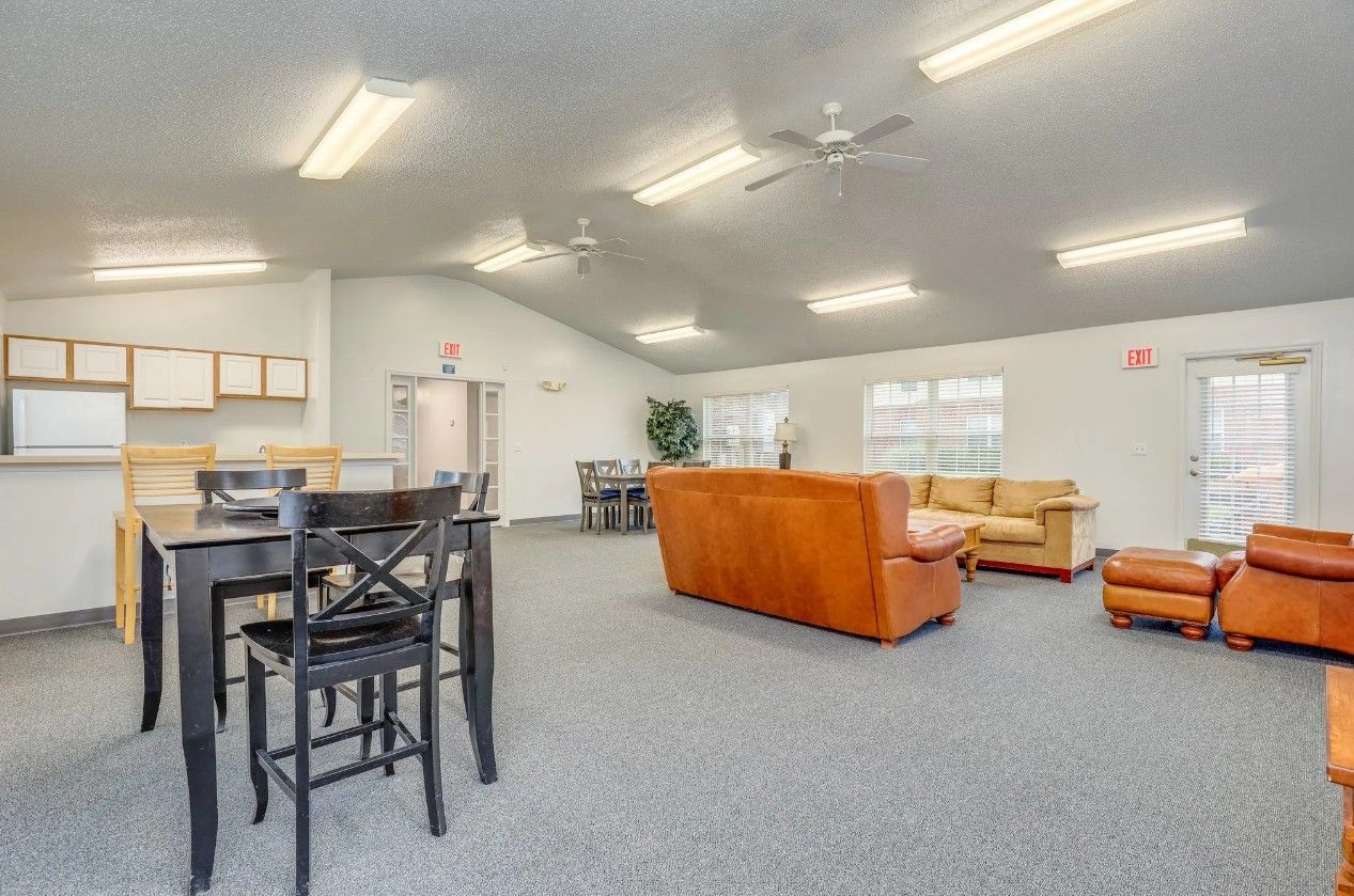 A spacious, carpeted community room with a dining set, orange sofas, and a counter area under bright overhead lighting.