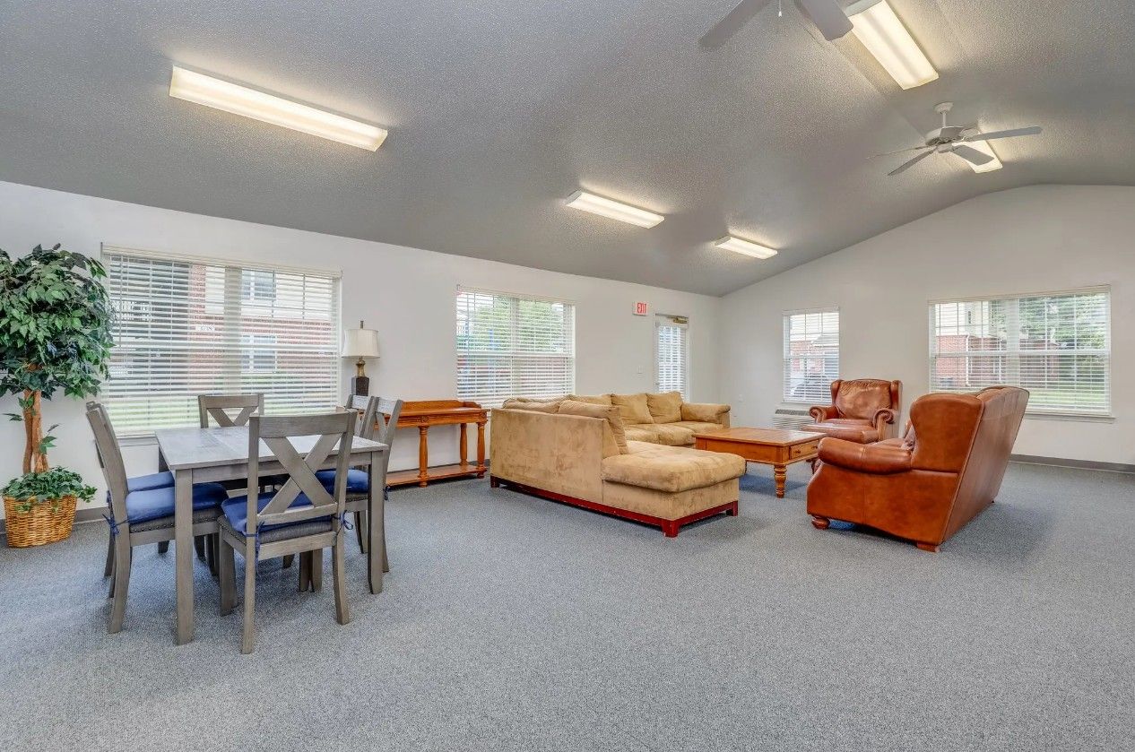 A bright, open community room with gray carpet, a dining table, a beige sectional sofa, and a leather armchair.