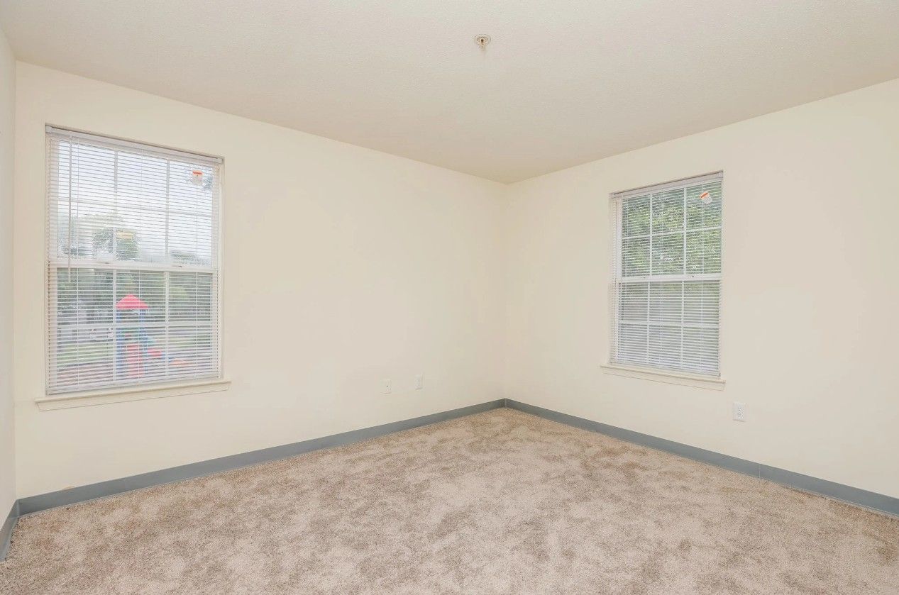 An empty bedroom with cream-colored walls, beige carpet, and two windows with white blinds.