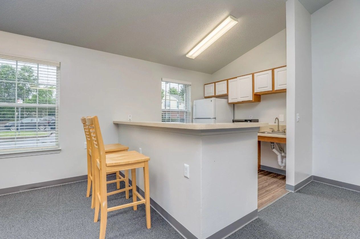A kitchen interior with a breakfast bar, two wooden bar stools, white cabinets, a refrigerator, and a window.