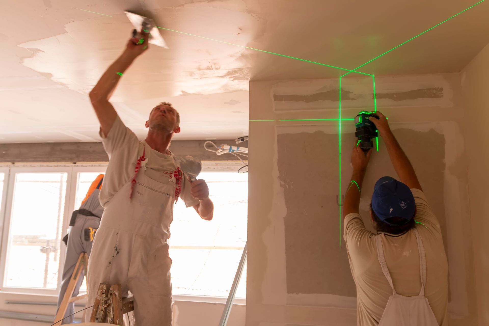 A man is using a laser to measure a wall while another man is plastering the ceiling.