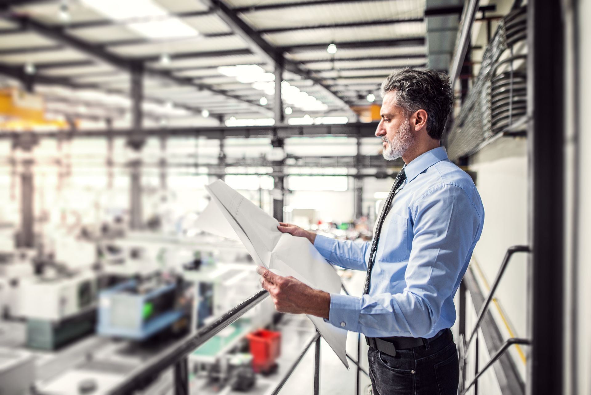 A man is standing on a balcony in a factory looking at a blueprint.