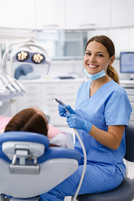 Dentist in blue scrubs smiling beside a patient in a dental chair, holding a dental tool in a bright clinic