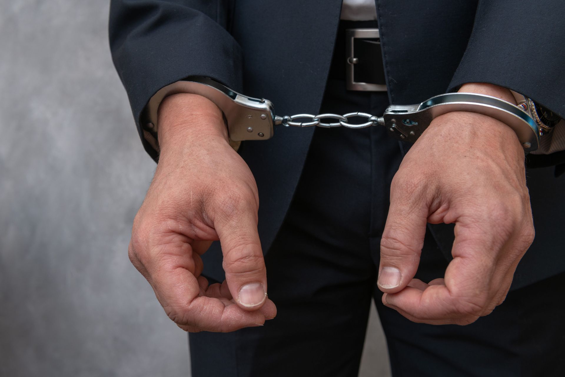 Hands in metal handcuffs against a dark suit and belt.
