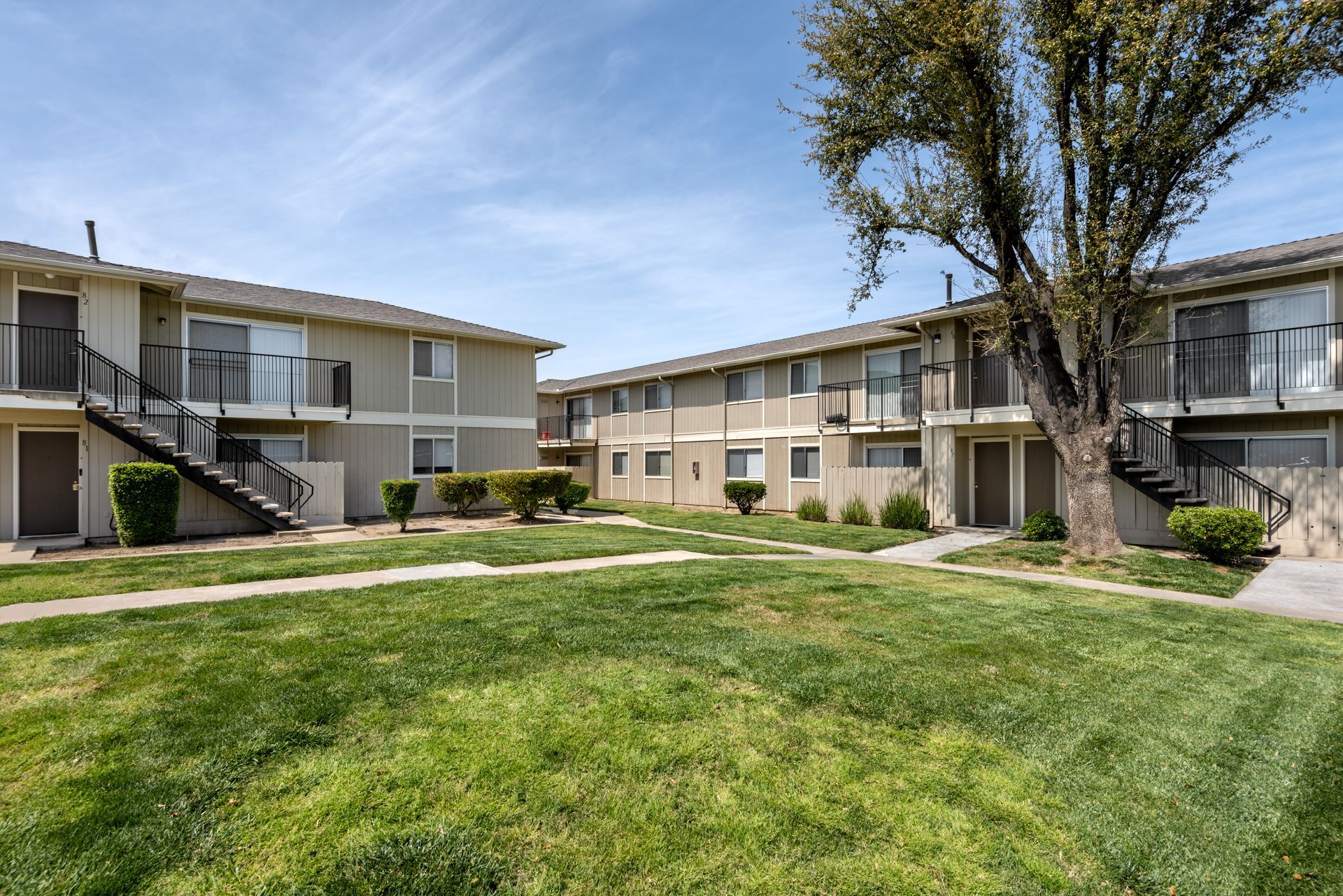 Apartment complex with two-story buildings, balconies, and a grassy courtyard on a sunny day.