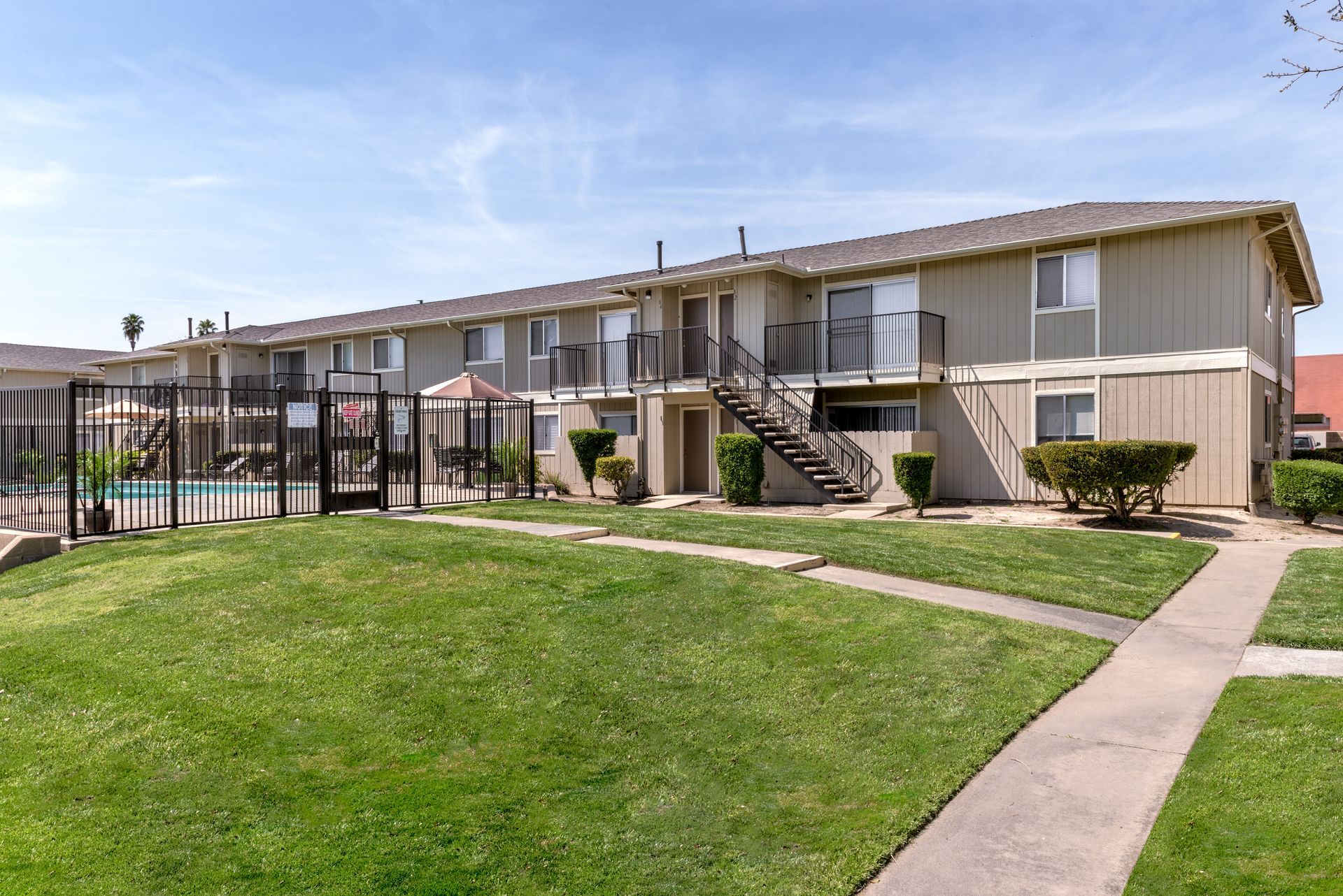 Apartment building with a pool behind a fence, sunny day. Green lawn and walkways.