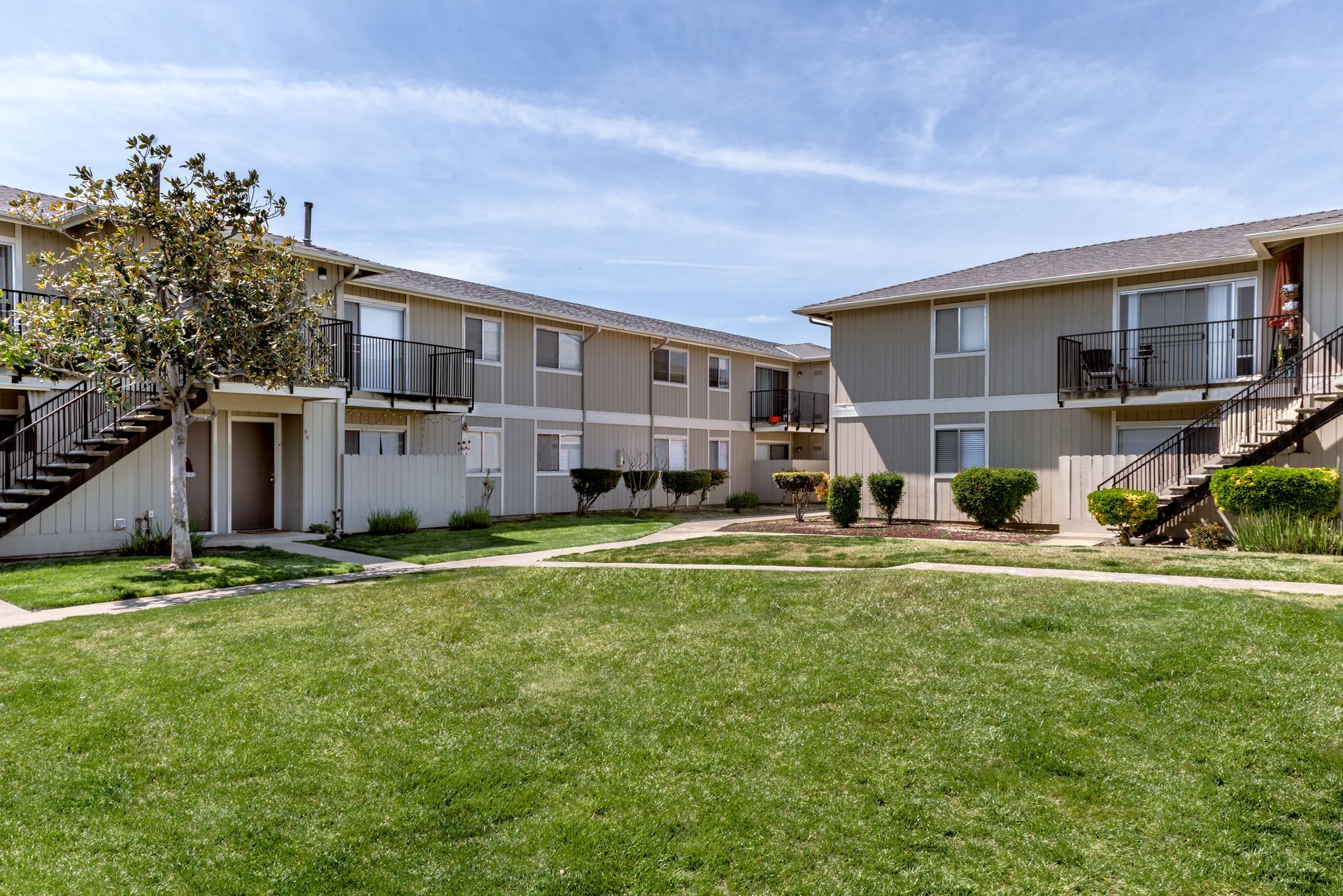 Two-story apartment buildings with balconies face each other over a grassy courtyard under a blue sky.
