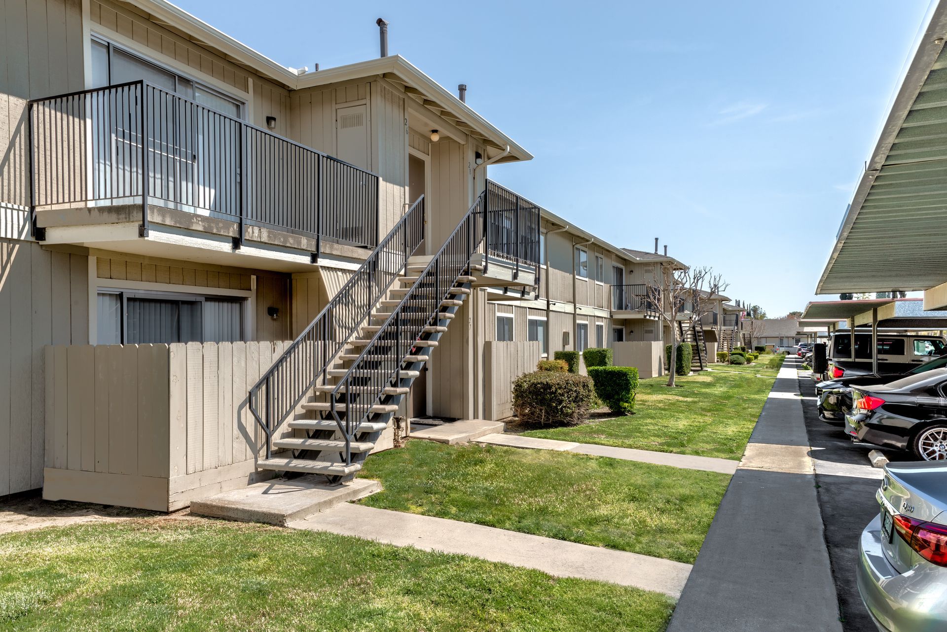 Apartment building with stairs to upper units; cars parked in covered spaces along a grassy path.