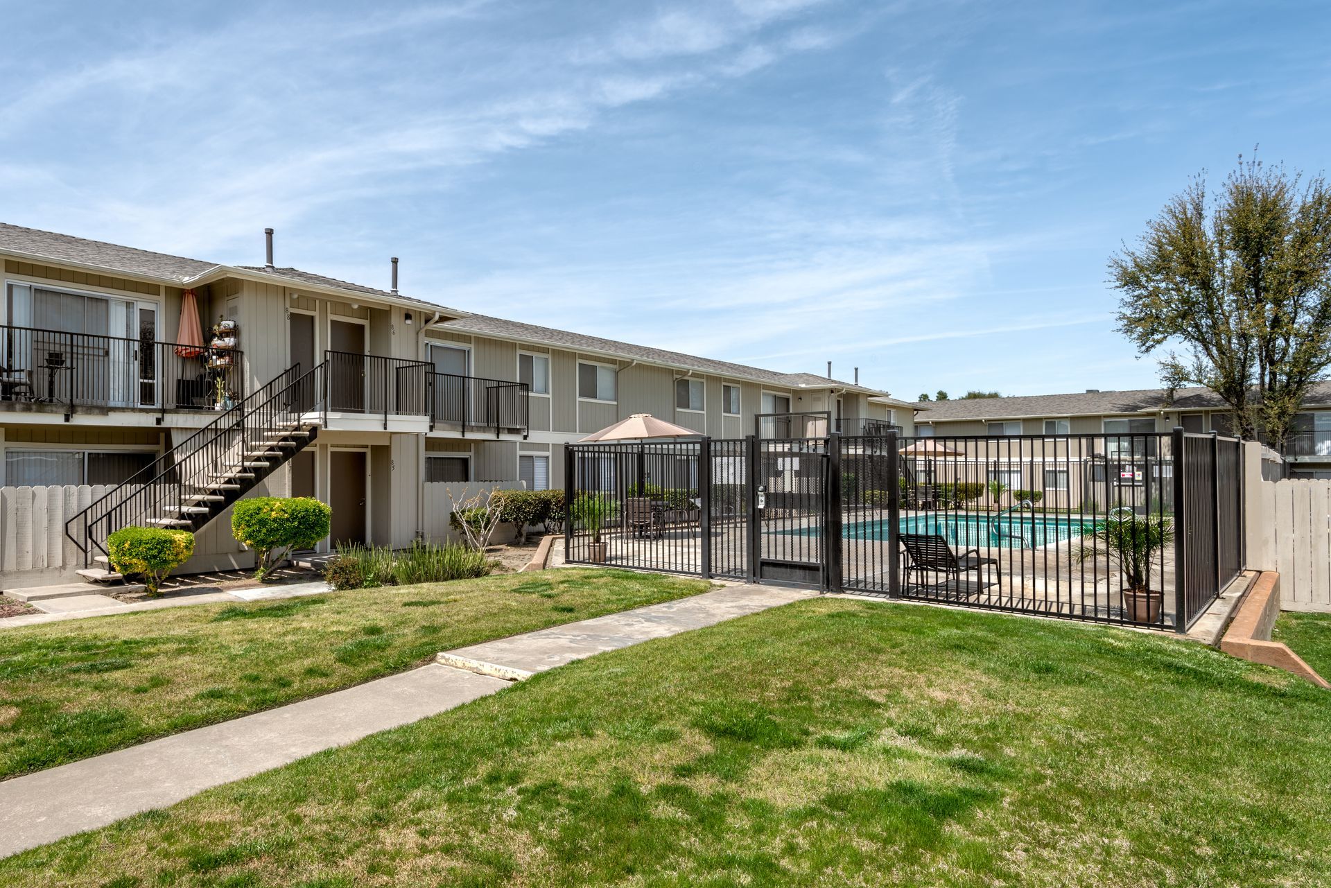 Apartment complex with a pool, grassy lawn, and fence on a sunny day.
