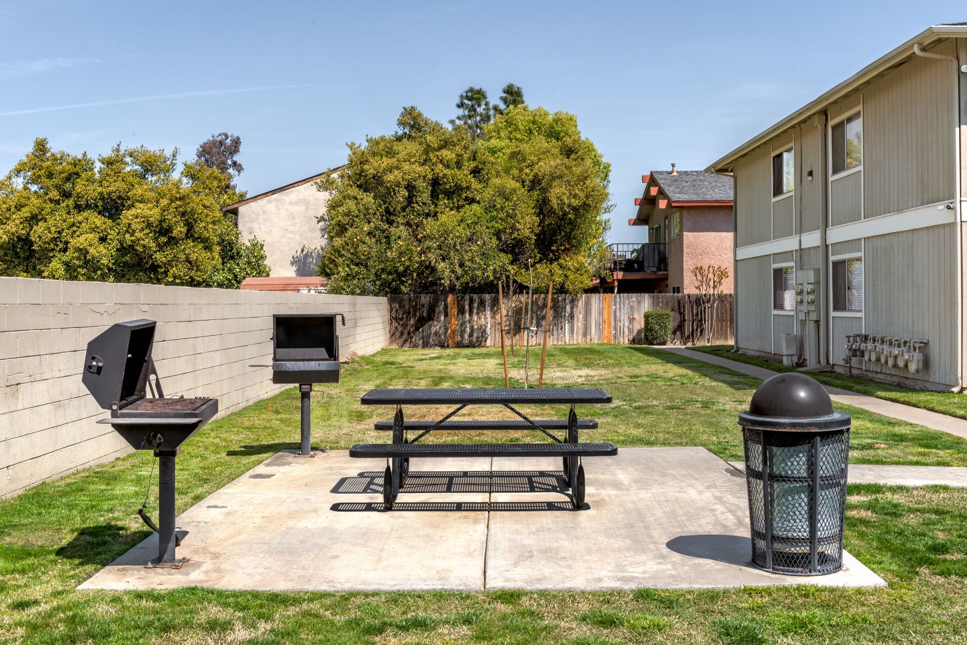 Outdoor picnic area with grill, table, and trash can near apartment buildings.