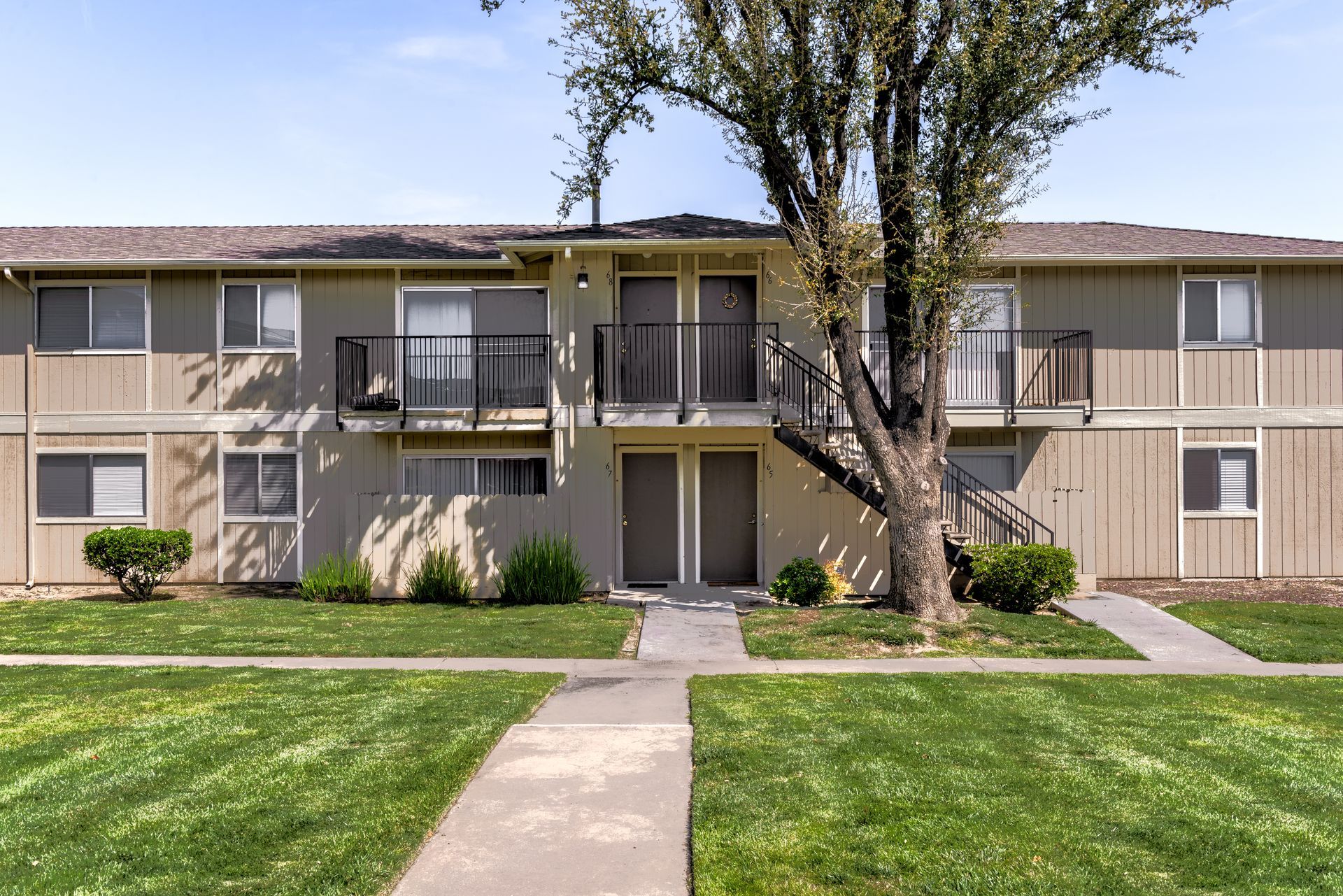 Two-story beige apartment building with balconies, grass, and a tree in the foreground.