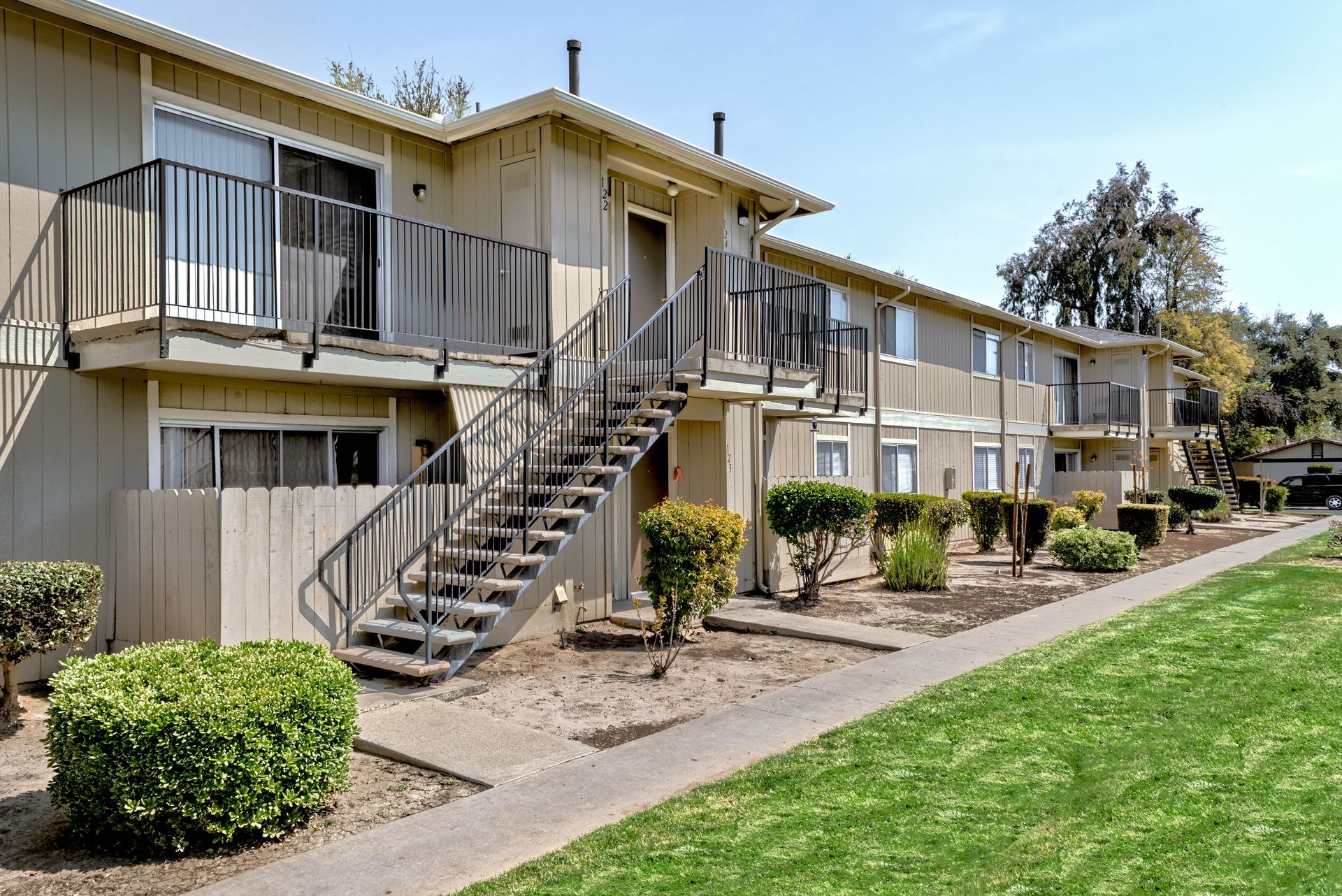 Apartment building with beige siding, black stairways, and green landscaping on a sunny day.
