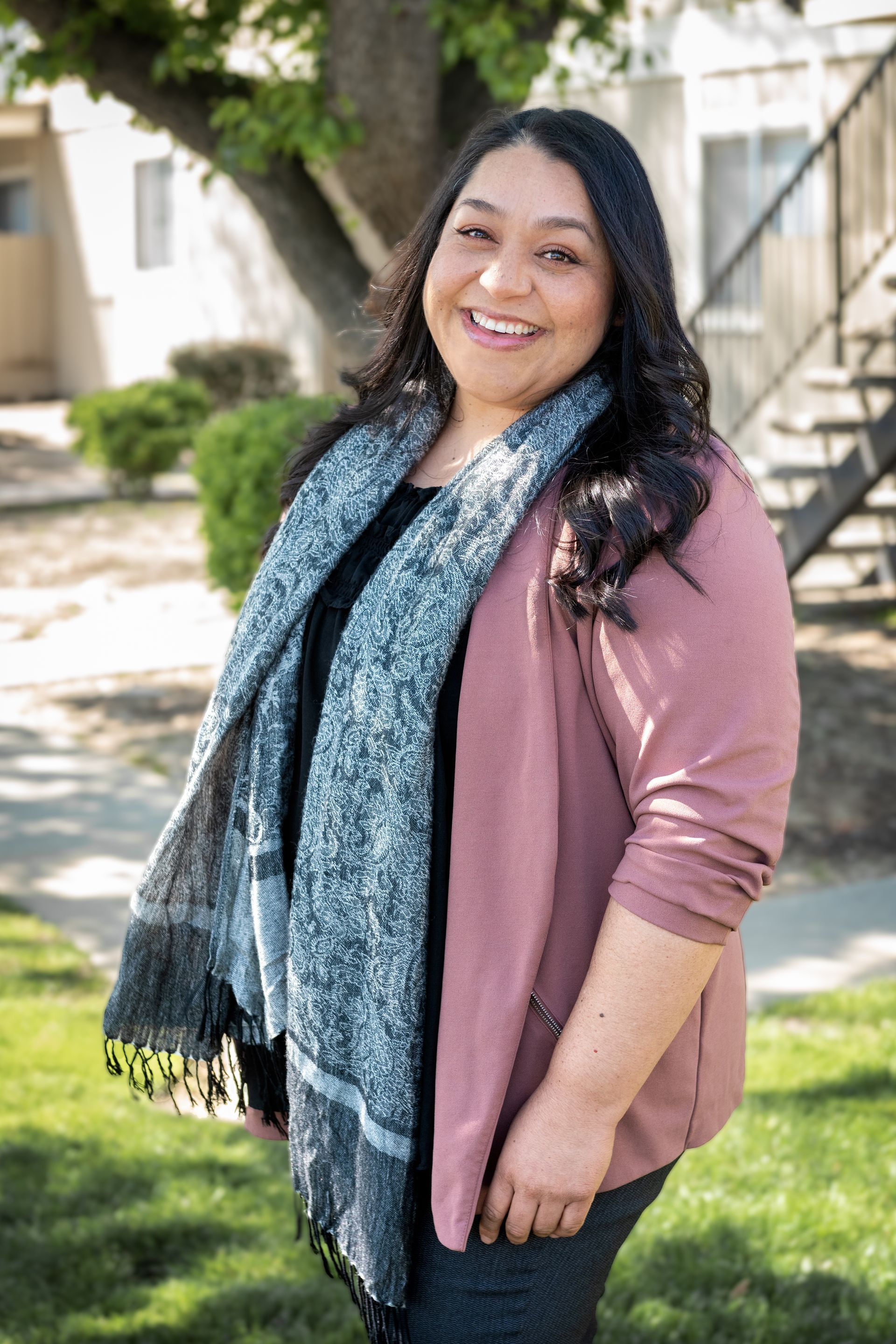 Woman in a pink cardigan and patterned scarf smiles outdoors in front of a tree and stairs.