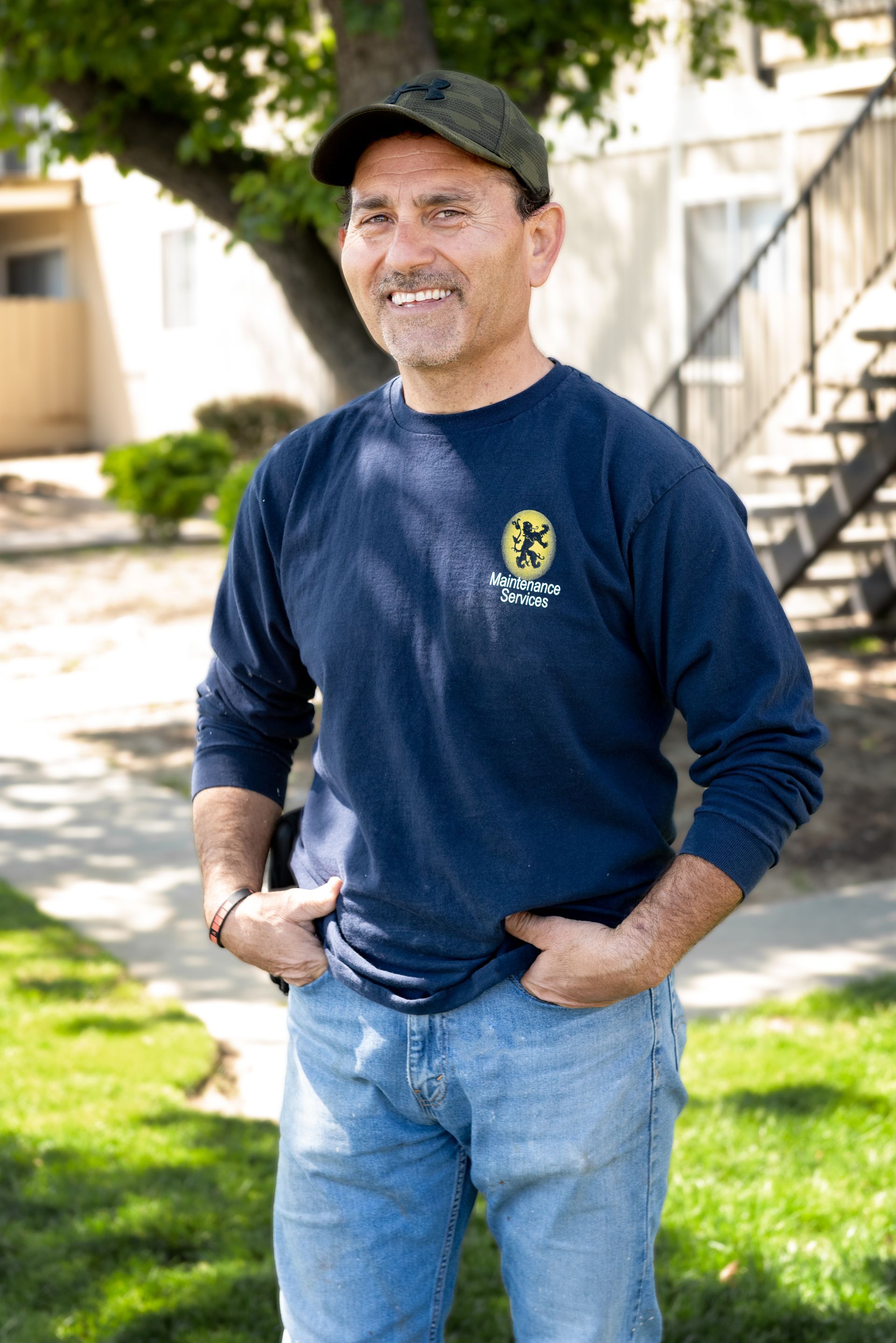 Man in blue shirt and jeans stands outside, hands in pockets, smiles.