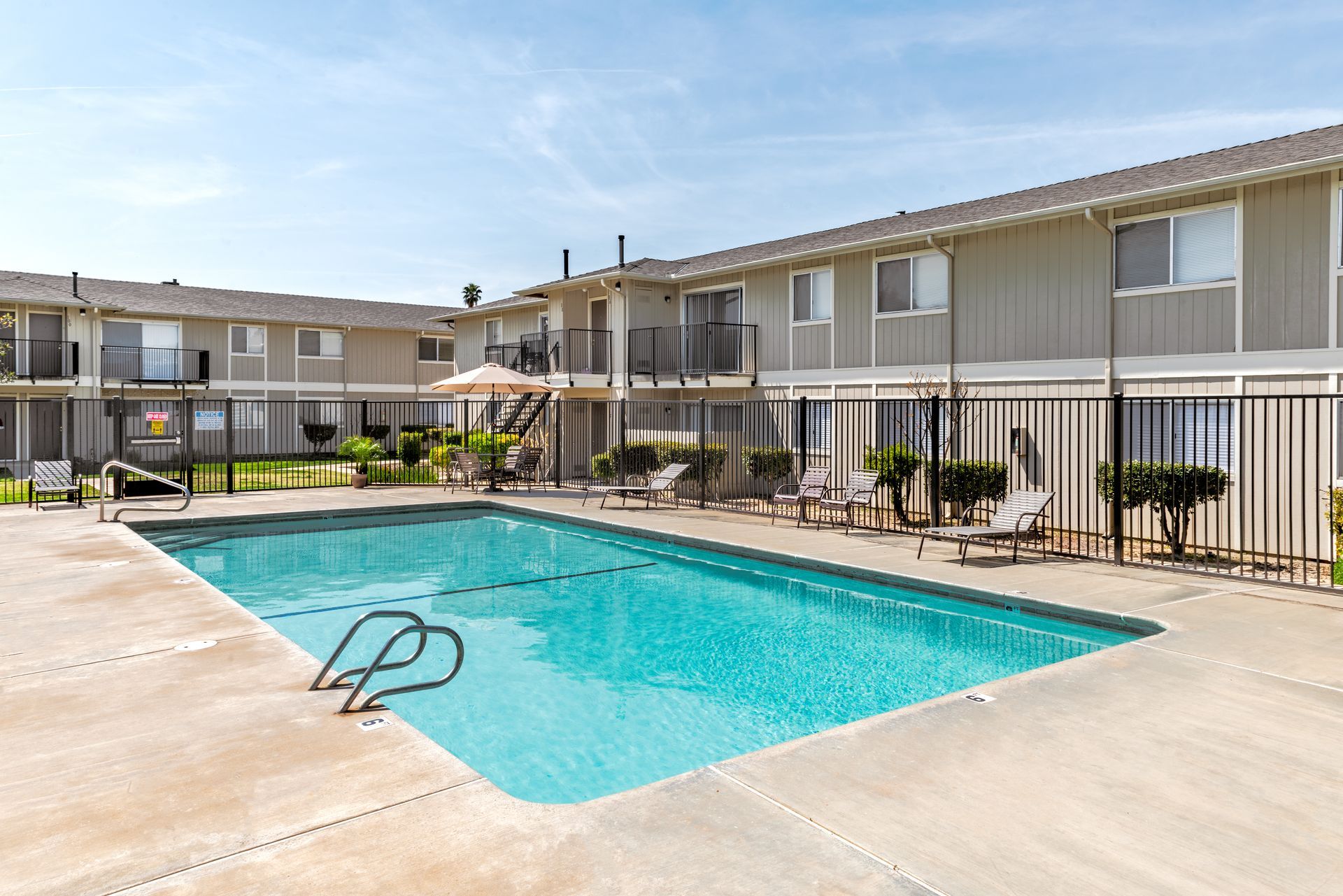 Apartment complex with a blue pool, lounge chairs, and beige buildings under a sunny sky.