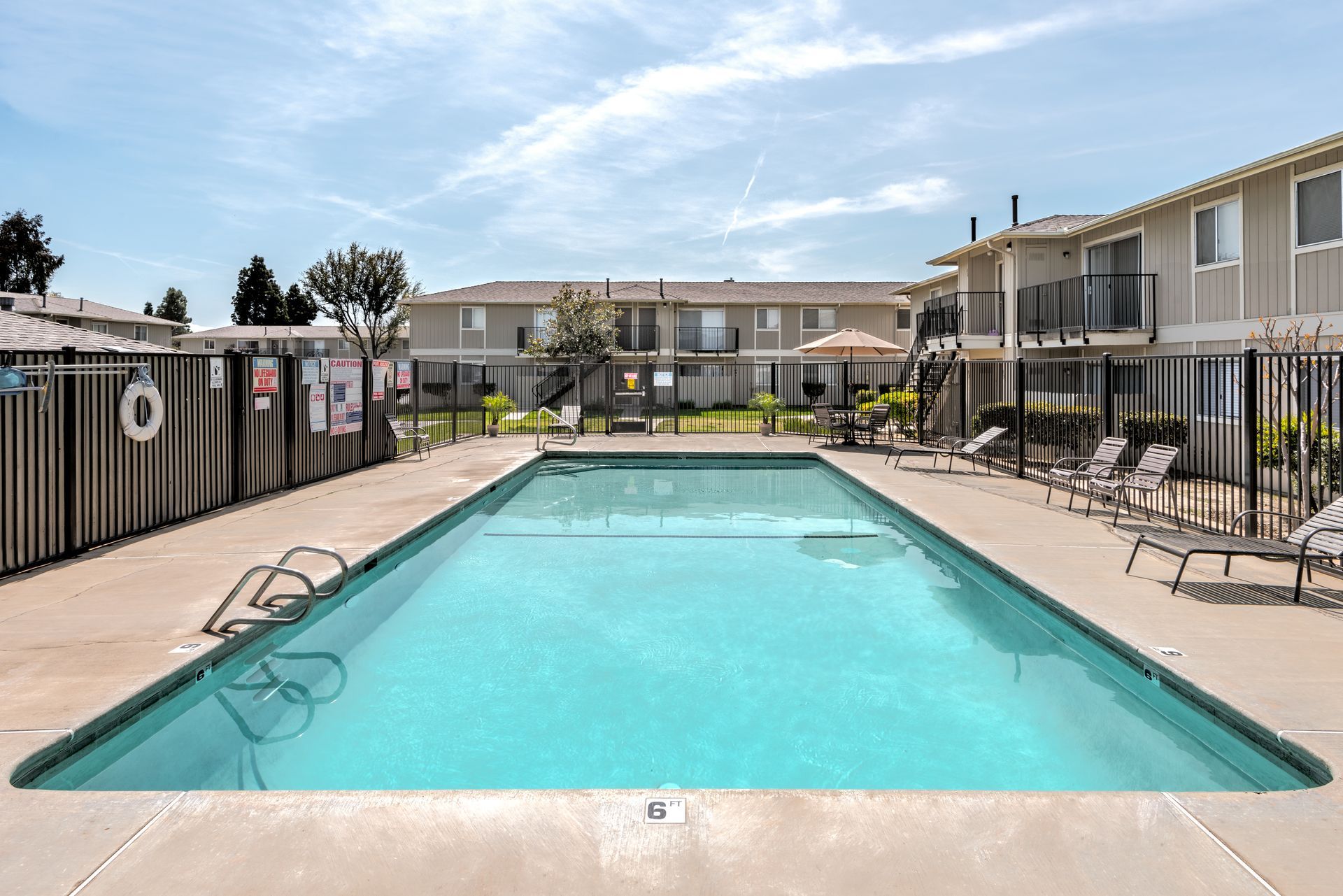 Swimming pool in front of an apartment complex on a sunny day with lounge chairs and a fence.