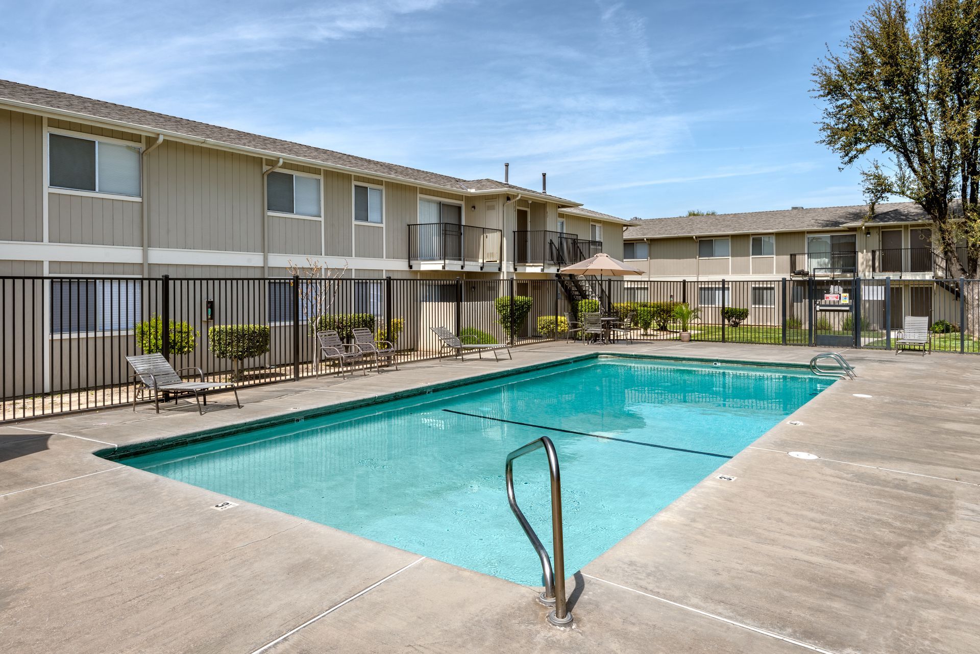 Apartment complex with a swimming pool, a fence, and outdoor seating under a blue sky.