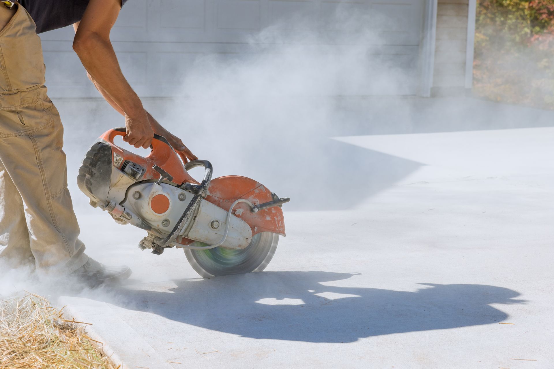 Worker performing industrial concrete cutting with walk behind saw on dusty slab outdoors area.