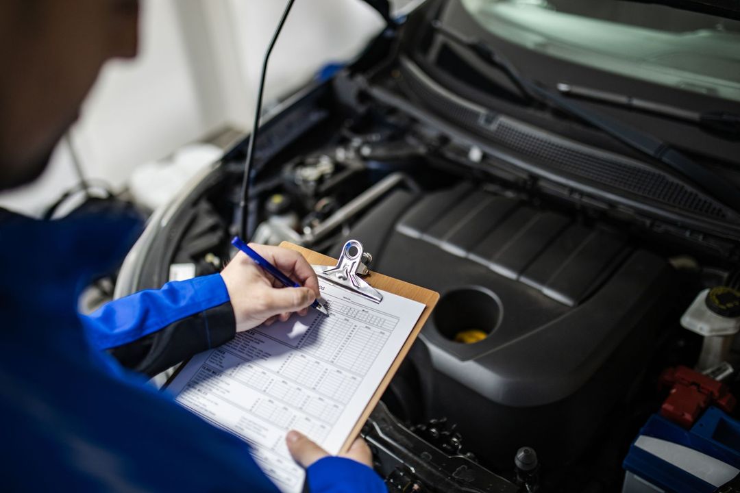 A mechanic is writing on a clipboard while looking under the hood of a car.