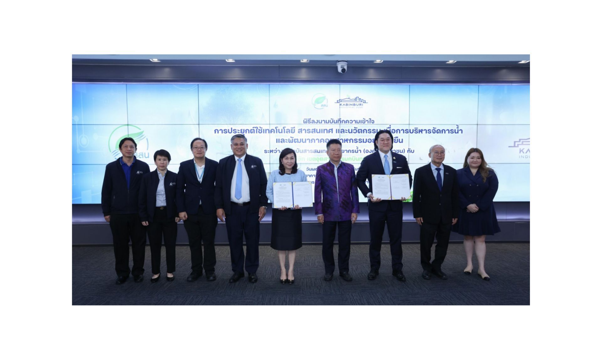 A group of people are standing in front of a large screen holding papers.