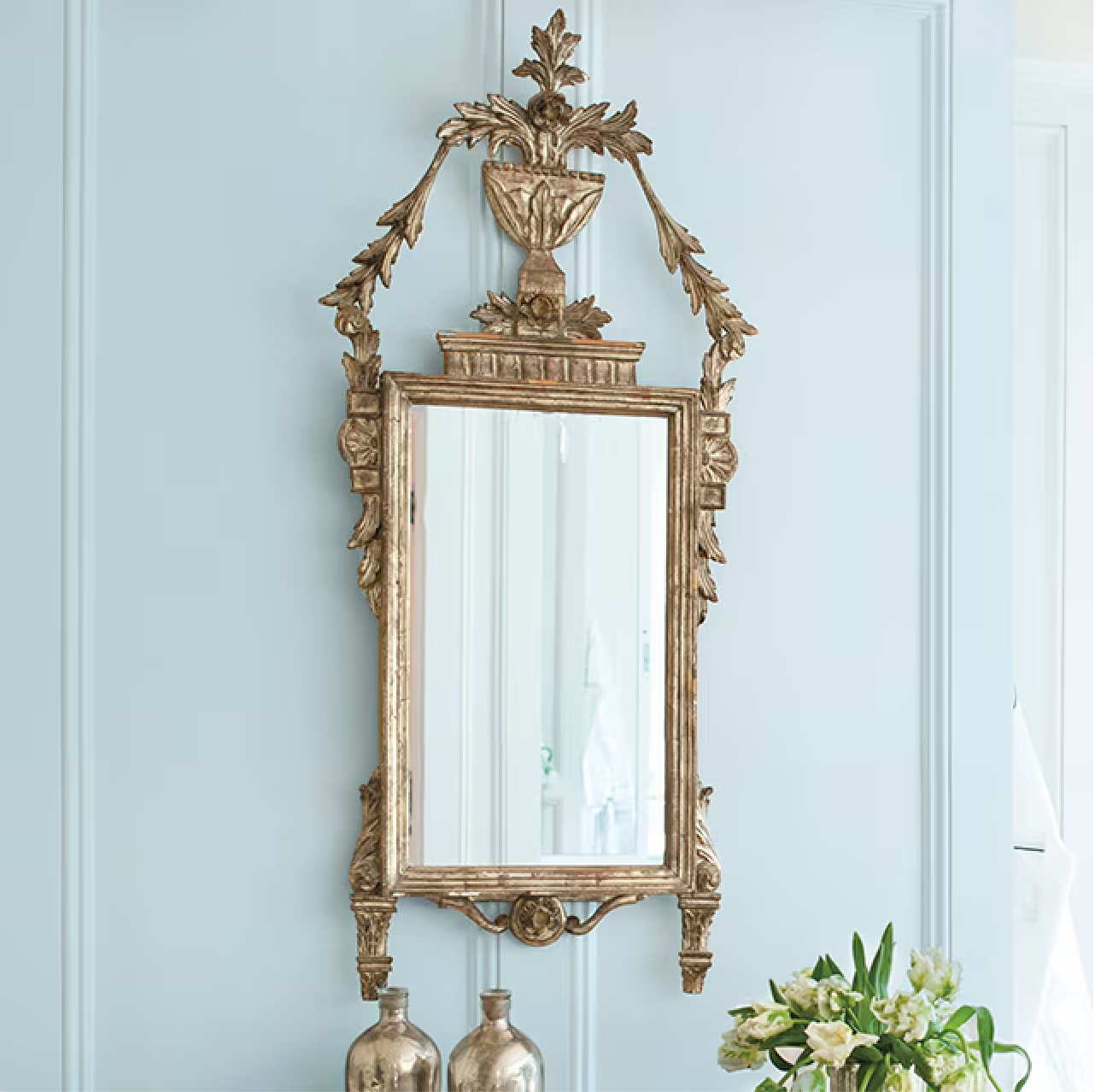 Ornate gold mirror hanging on a pale blue wall with decorative leaves and a vase detail.