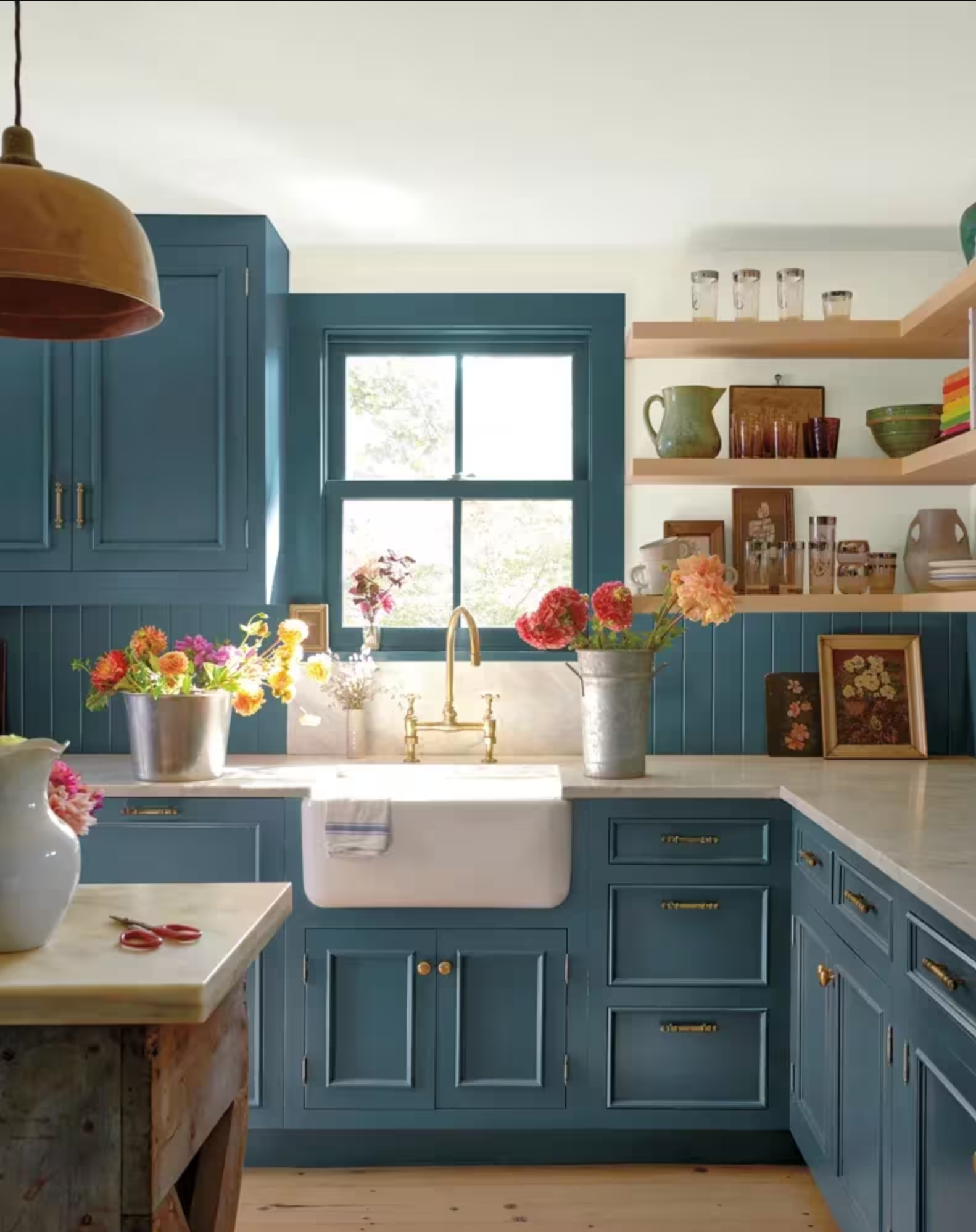 Blue kitchen with white sink, wooden shelves with decor, and colorful flowers.