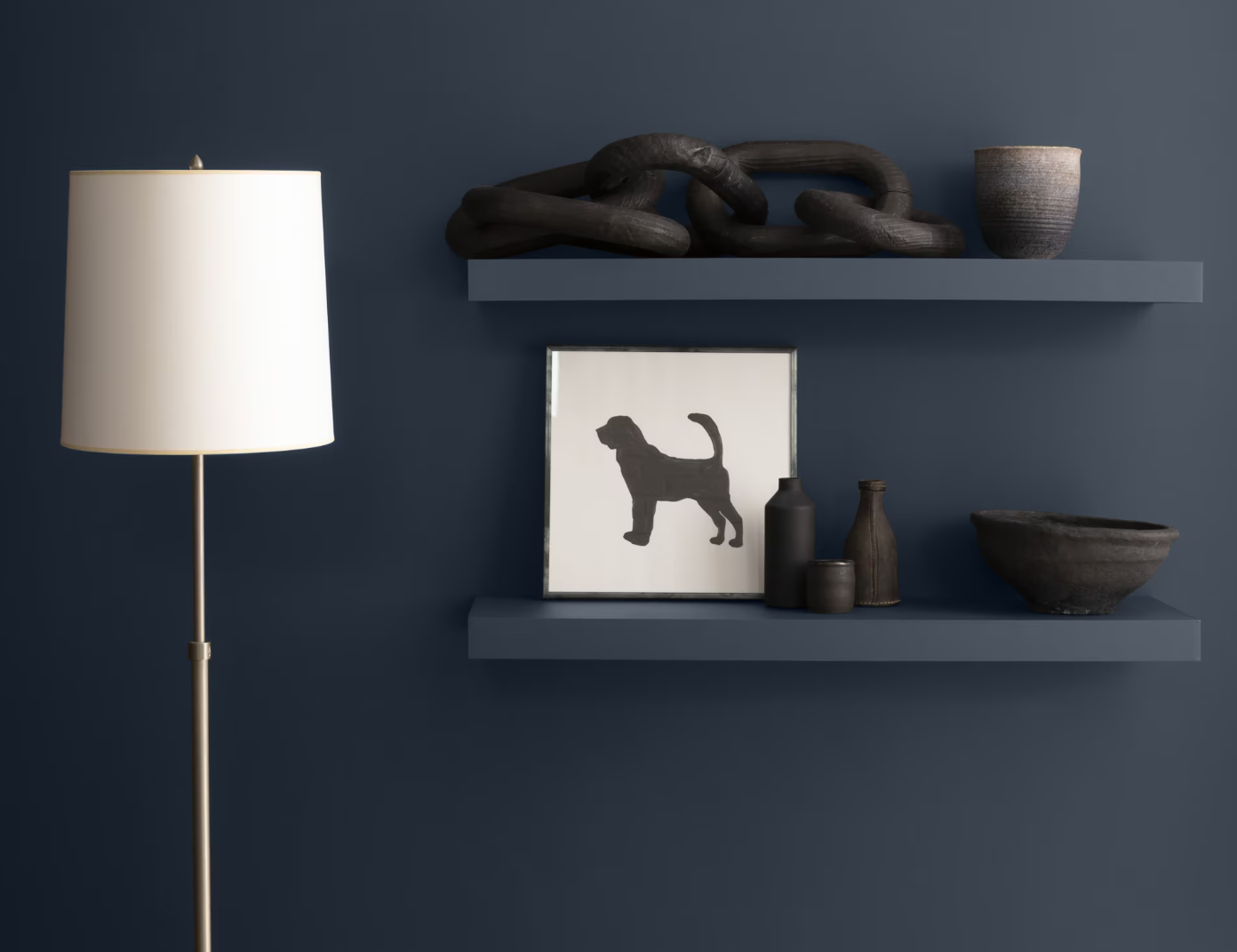 A floor lamp stands next to a wall with two dark floating shelves displaying a framed dog silhouette and minimalist decor.
