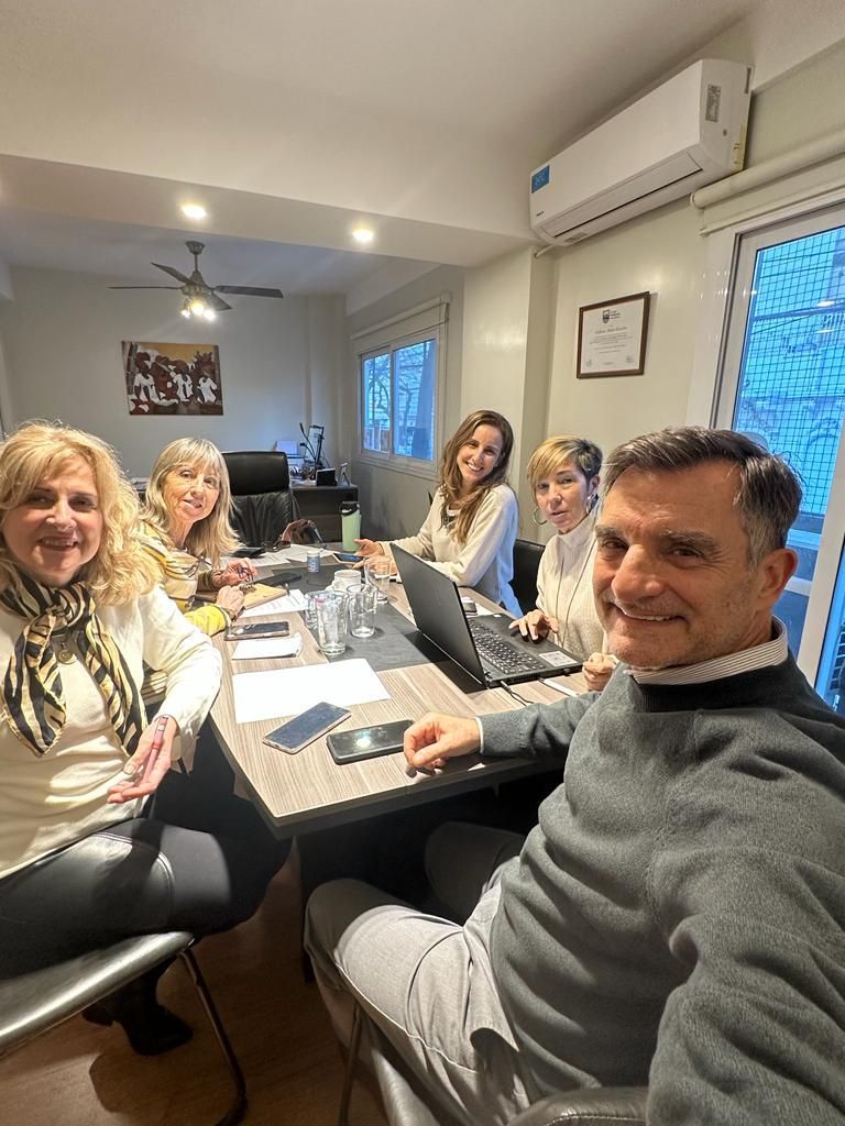 Cinco personas sentadas alrededor de una mesa, una de ellas tomándose una selfi. Oficina bien iluminada.