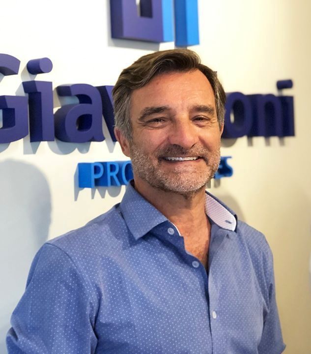 Un hombre sonriente con una camisa azul abotonada está frente a un logotipo azul en una pared blanca.