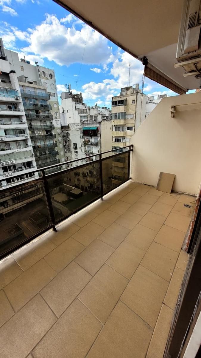 Edificio alto de apartamentos con balcones, peatones en la acera, coche rojo aparcado en la calle, cielo azul.