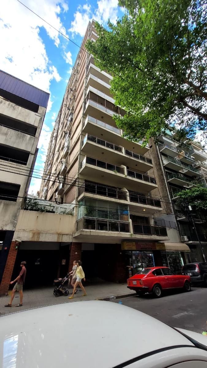 Edificio alto de apartamentos con balcones, peatones en la acera, coche rojo aparcado en la calle, cielo azul.