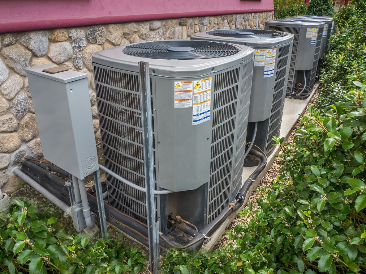 Outdoor HVAC condenser units beside a building wall, surrounded by shrubs and stone siding