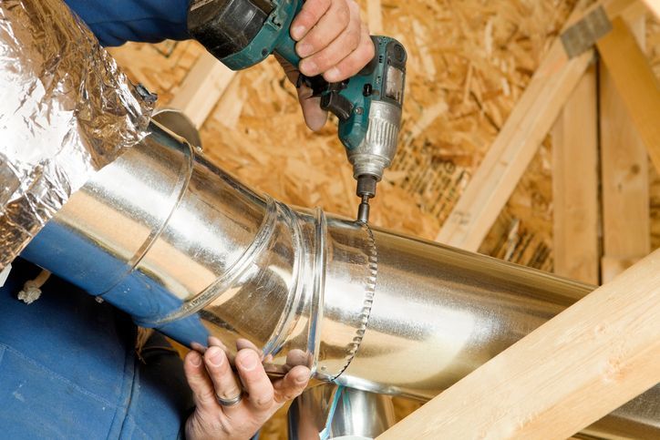 Technician using a drill to install a metal HVAC duct in an attic with wooden framing