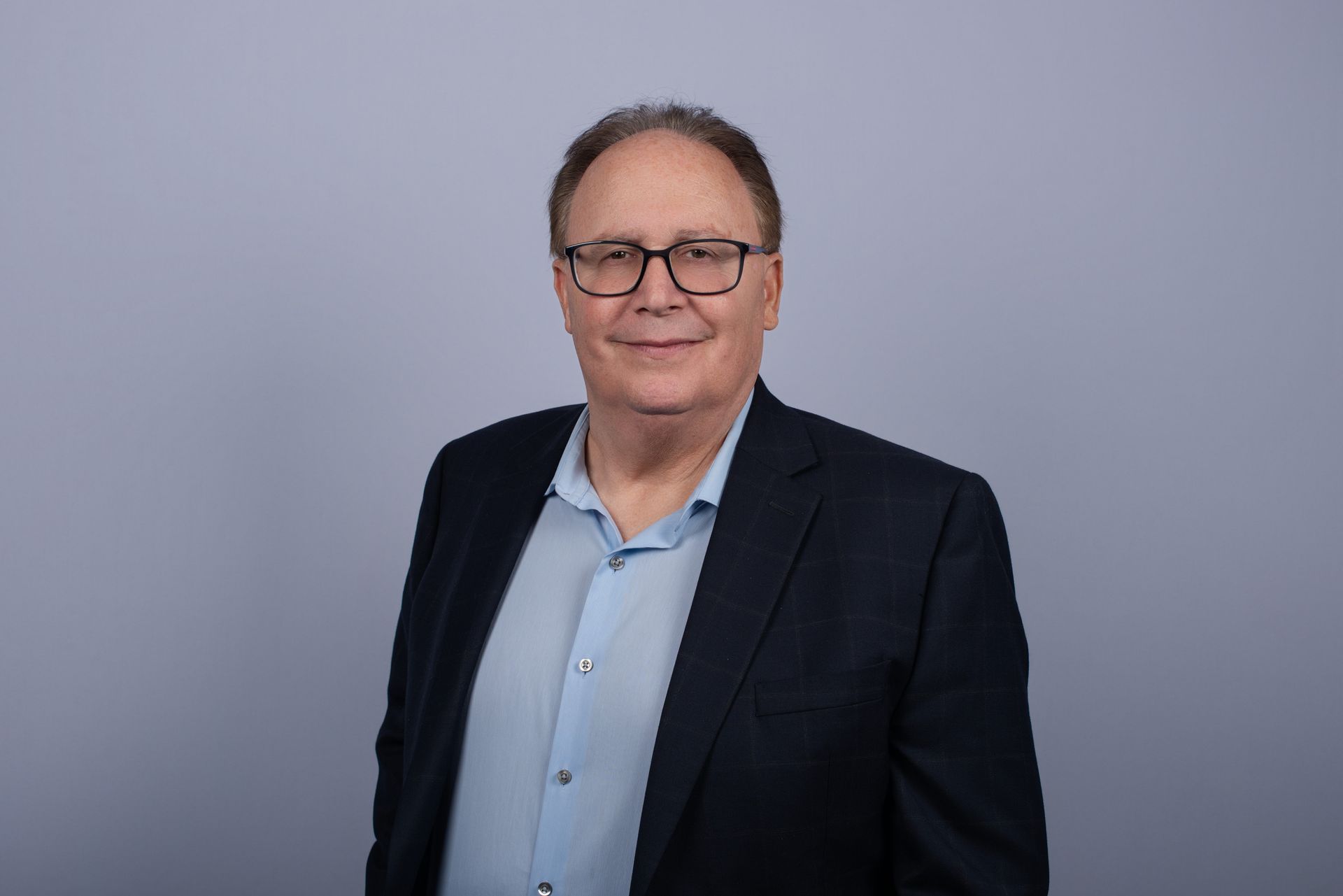 A man with glasses smiles, wearing a light blue button-down shirt, against a light blue background.