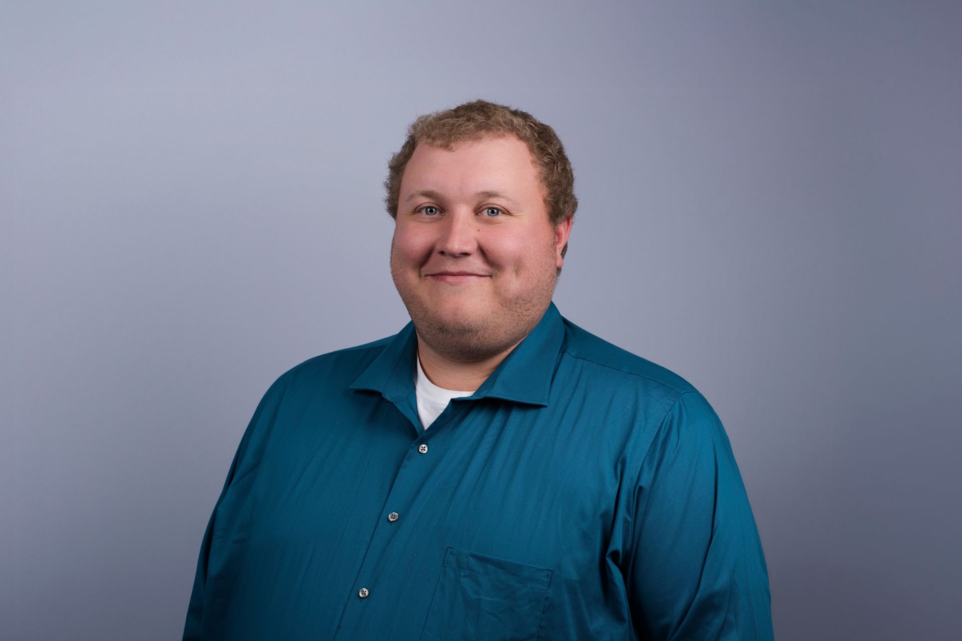 Man with a short, spiky haircut and stubble, smiling. Wearing a blazer and light blue shirt, with a blue background.