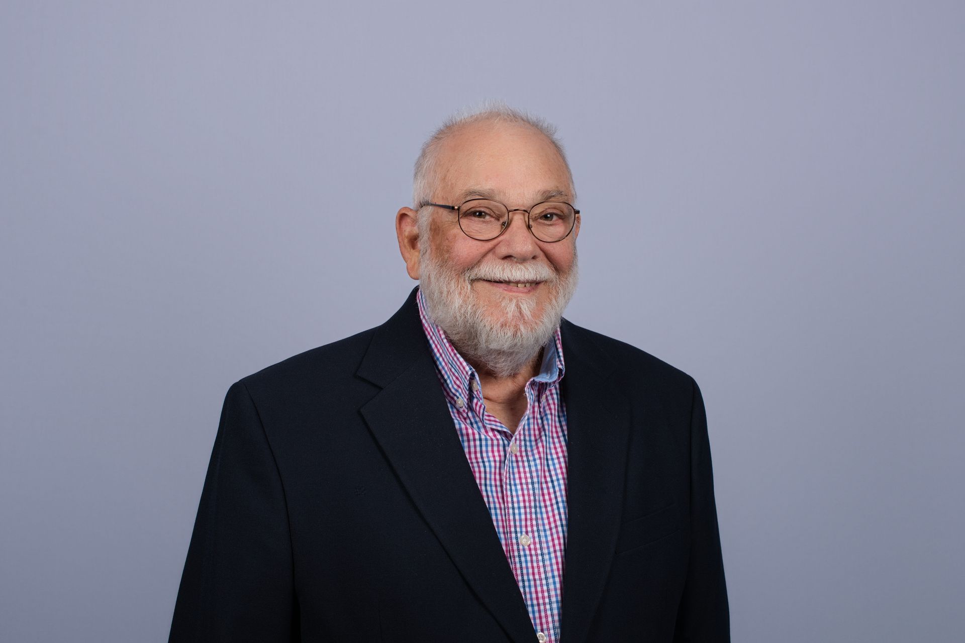Man with a short, spiky haircut and stubble, smiling. Wearing a blazer and light blue shirt, with a blue background.