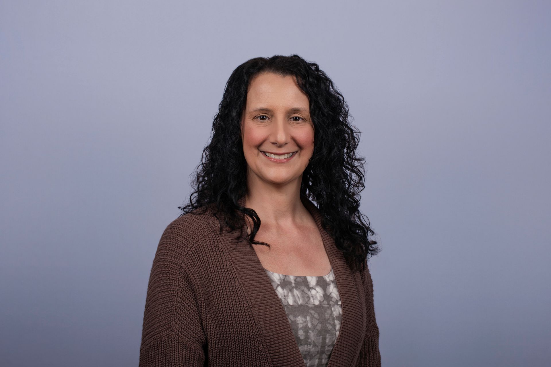 Man with a short, spiky haircut and stubble, smiling. Wearing a blazer and light blue shirt, with a blue background.