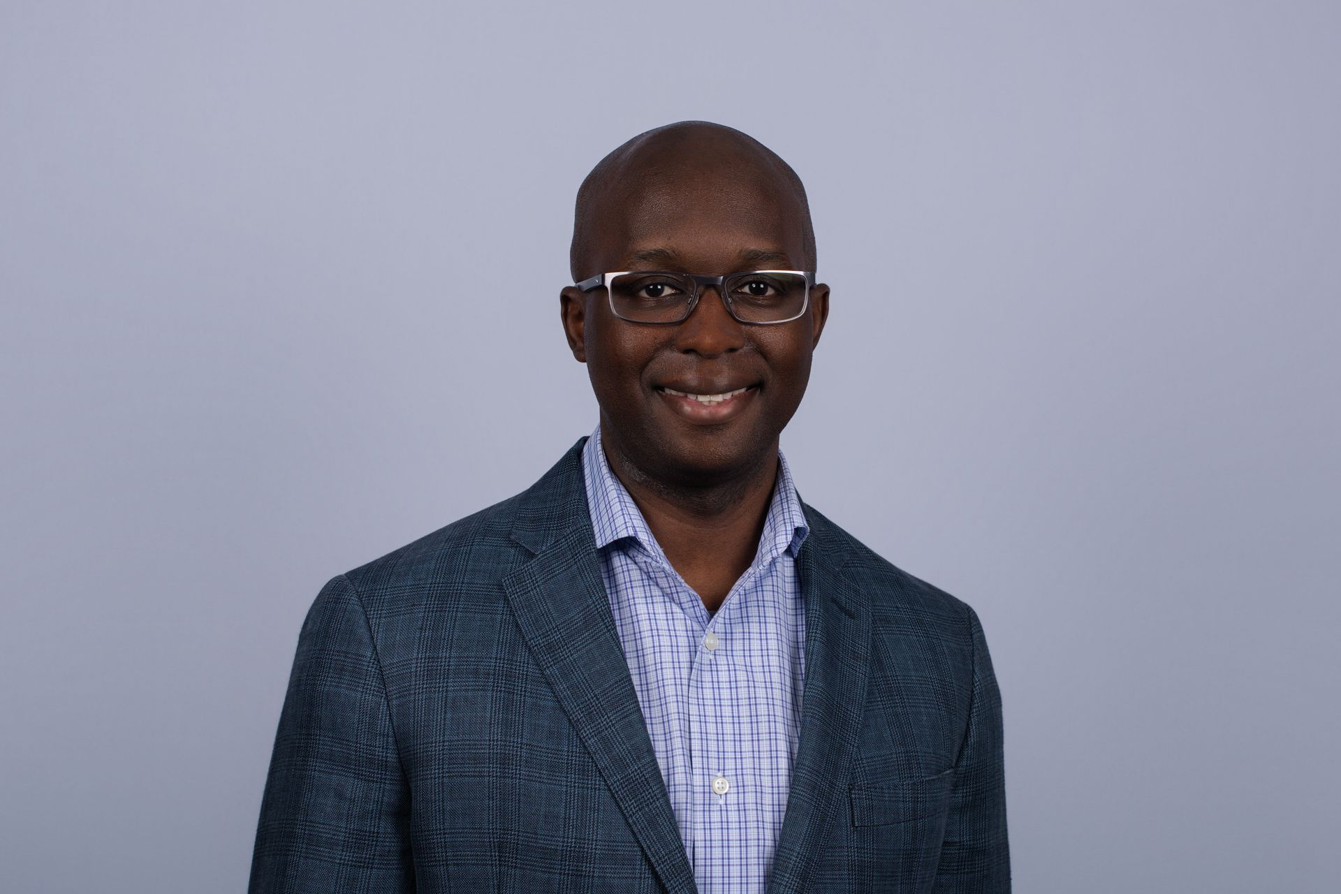 A man with glasses smiles, wearing a light blue button-down shirt, against a light blue background.