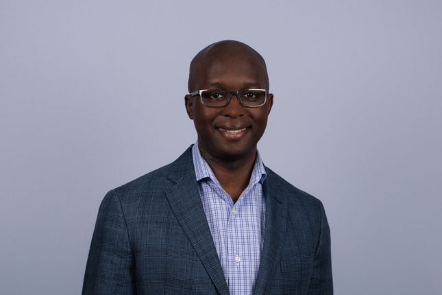 A man with glasses smiles, wearing a light blue button-down shirt, against a light blue background.