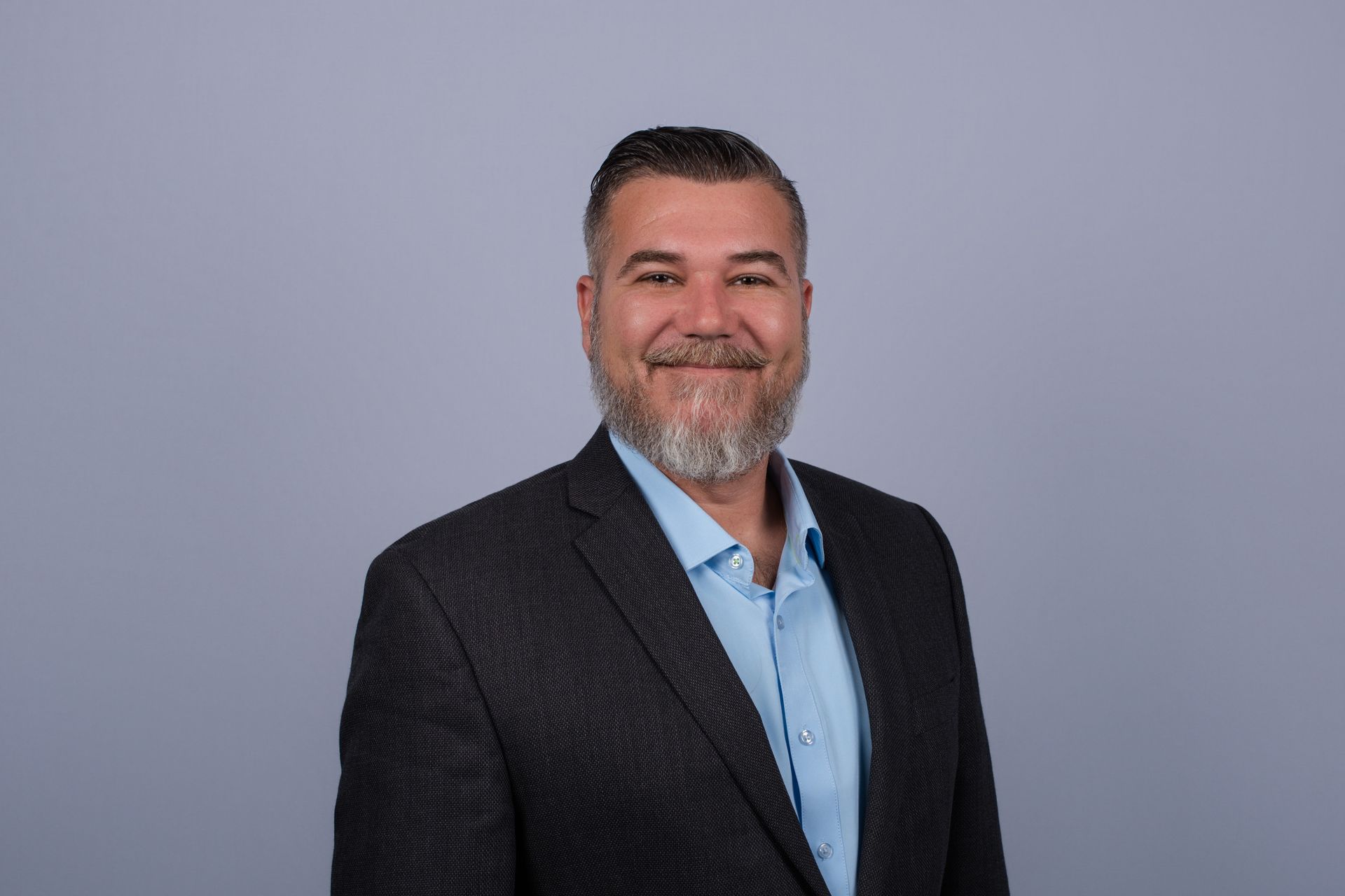 Man with a short, spiky haircut and stubble, smiling. Wearing a blazer and light blue shirt, with a blue background.
